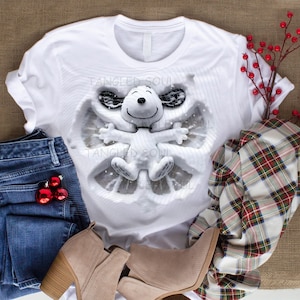 Könnte beinhalten: Weißes T-Shirt mit einem Snoopy-Design als Schneeengel in Grau- und Weißtönen. Das Bild zeigt auch blaue Jeans, rote Weihnachtskugeln und beige Stiefeletten. Ein kariertes Hemd und rote Beeren ergänzen die festliche Szene.