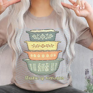 Może przedstawiać: Beżowa koszulka z nadrukiem przedstawiającym stos czterech retro naczyń do zapiekania w odcieniach niebieskiego, żółtego i pomarańczowego. Naczynia mają kwiatowe i geometryczne wzory. Pod spodem znajduje się napis "Stackin up memories".