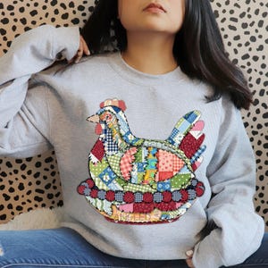 Könnte beinhalten: Hellgraues Sweatshirt mit einem farbenfrohen Patchwork-Hühnerdesign. Das Huhn besteht aus verschiedenen Stoffmustern und -farben, darunter Rot, Blau, Grün und Rosa. Das Wort "ANGLED" ist unten sichtbar.