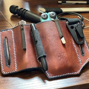 Könnte beinhalten: Ein braunes Lederetui mit Nähten. Das Etui hat drei Fächer zum Tragen eines Stifts, eines Multitools und eines kleinen Messers. Das Multitool hat einen türkisfarbenen Griff und der Text "Leatherman" ist auf dem Werkzeug sichtbar.