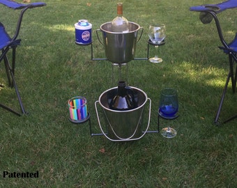 May include: A black metal wine and beverage stand with two stainless steel buckets, each holding a bottle. The stand has holders for glasses and a can. The stand is on a grassy lawn with two blue folding chairs.