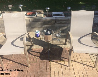 May include: Two gray mesh patio chairs with a metal ice bucket and a glass on a tripod stand in the middle. The ice bucket has a glass bottle inside. There is a can of beverage next to the ice bucket. The chairs are on a brick patio.