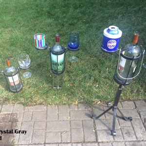 May include: Three black metal wine bottle holders with a tripod base. Each holder has a space for a bottle and two glasses. The holder on the right has a space for a can cooler. The holders are on a brick patio with green grass in the background. Color: Crystal Gray. Patented.