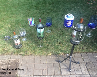 May include: Three black metal wine bottle holders with a tripod base. Each holder has a space for a bottle and two glasses. The holder on the right has a space for a can cooler. The holders are on a brick patio with green grass in the background. Color: Crystal Gray. Patented.