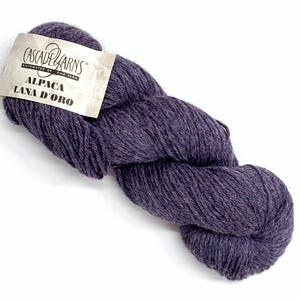 May include: A skein of dark purple Cascade Yarns Alpaca Lana D'Oro yarn. The yarn is a deep, rich shade of purple, with a soft, textured appearance. The label reads "Cascade Yarns" and "Alpaca Lana D'Oro".