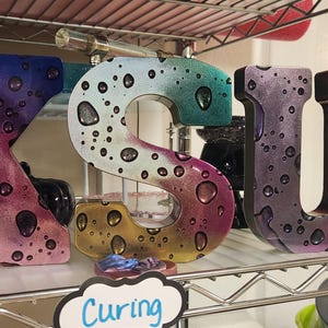 May include: Wooden letters spelling "KSU" with a rainbow and water drop design. The letters are on a metal shelf.