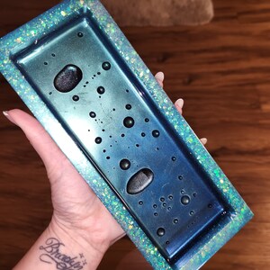 May include: A teal and blue rectangular tray with a glittery border. The tray has a clear resin surface with black air bubbles.