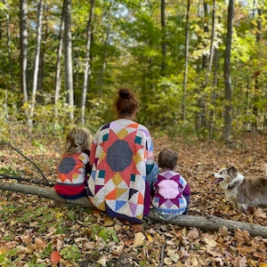 May include: A woman and two children wearing colourful, crocheted sweaters with starburst designs, sitting on a log in a forest. A dog stands nearby. The sweaters feature geometric patterns in various colours.