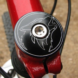 May include: Black bicycle stem cap with a silver wolf head design. The cap has a silver hex bolt in the center.