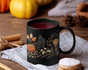 May include: A black mug with a floral design featuring fall colors, filled with a dark red beverage. The mug is sitting on a gray linen napkin with a small, round cookie dusted with powdered sugar.
