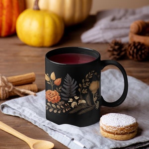 May include: A black mug with a floral design featuring fall colors, filled with a dark red beverage. The mug is sitting on a gray linen napkin with a small, round cookie dusted with powdered sugar.