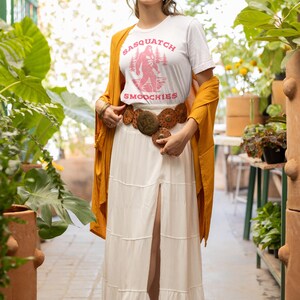Peut inclure: Une femme portant un t-shirt blanc avec un graphique rose d'un sasquatch tenant une tasse qui dit "Sasquatch Smoothies". Elle porte un chapeau marron, une jupe maxi blanche et un cardigan jaune moutarde.