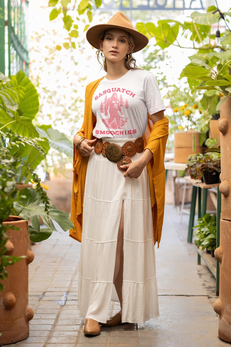 Peut inclure: Une femme portant un t-shirt blanc avec un graphique rose d'un sasquatch tenant une tasse qui dit "Sasquatch Smoothies". Elle porte un chapeau marron, une jupe maxi blanche et un cardigan jaune moutarde.