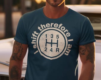 Pode incluir: Uma camiseta azul marinho com um gráfico branco de uma alavanca de câmbio de carro e o texto "I shift therefore I am". O gráfico inclui os números 1, 3, 5, 2, 4 e R.