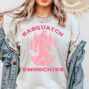 Peut inclure: Un sweat-shirt gris clair avec un graphique rose d'un sasquatch tenant une tasse avec le texte "Sasquatch Smoochies".