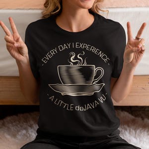 Könnte beinhalten: Schwarzes T-Shirt mit einer weißen Grafik einer Kaffeetasse und Untertasse, aus der Dampf aufsteigt. Der Text lautet: "- EVERY DAY I EXPERIENCE - A LITTLE deJAVA VU -."