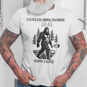 May include: White t-shirt with a black and white illustration of Bigfoot wearing bunny ears and holding Easter eggs. The text "Easter Egg Hiding Champion" and "Happy Easter" is printed on the shirt.