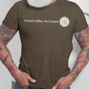 以下が含まれることがあります： 「Grind Coffee, Not Gears」の文字と白いギアシフトパターンのグラフィックが入った茶色のTシャツ。半袖で柔らかい素材です。