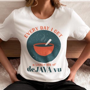May include: White t-shirt with a circular graphic featuring a steaming orange bowl and spoon. The text reads "EVERY DAY I GET" above the graphic and "a Little bit of deJAVA vu" below. The shirt is a casual, everyday wear item.