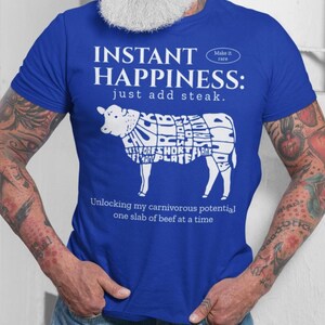 Op de afbeelding: Koningsblauw t-shirt met witte tekst en een diagram van een koe met verschillende vlees sneden. De tekst luidt "INSTANT HAPPINESS: just add steak" en "Unlocking my carnivorous potential one slab of beef at a time."