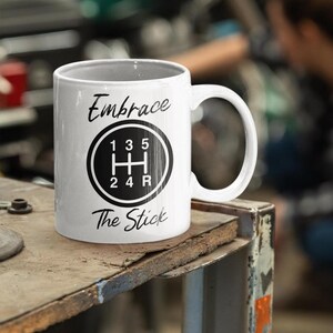 May include: White ceramic coffee mug with a black and white graphic of a stick shift gear pattern. The text "Embrace The Stick" is printed on the mug.