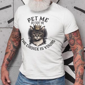 Könnte beinhalten: Weißes T-Shirt mit einer Grafik einer Katze mit Krone und dem Text "PET ME OR FIGHT ME THE CHOICE IS YOURS" in einem kreisförmigen Design. Das Design ist mittig auf dem T-Shirt platziert.