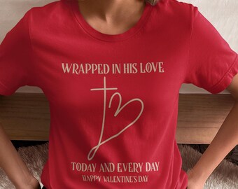May include: Red t-shirt with a white graphic design that says "Wrapped in His Love" with a cross and heart design. The text below says "Today and Every Day Happy Valentine's Day."