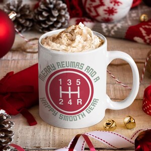 May include: White ceramic mug with a red and white gear shift design, filled with a whipped cream topped beverage. The mug has the text "Merry Reumas and a Happy Smooth Gear" around the gear shift graphic. Festive holiday theme.