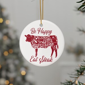 Op de afbeelding: Witte keramische kerstversiering met een rood silhouet van een koe met verschillende vleesdelen die zijn gelabeld. De tekst "Be Happy Eat Steak" is onder de koe gedrukt.