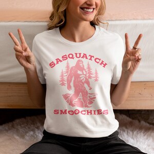 Peut inclure: T-shirt blanc avec un graphique rose d'un sasquatch tenant un smoothie. Le texte "Sasquatch Smoothies" est imprim&eacute; en rose sur le t-shirt.