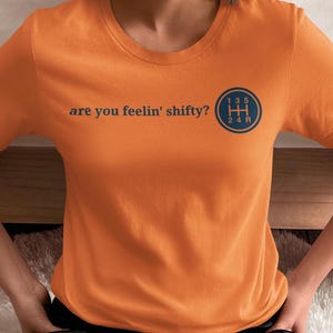 Könnte beinhalten: Orangefarbenes T-Shirt mit dem Text "are you feelin' shifty?" und einer blauen Grafik eines Schaltmusters. Das Shirt hat einen Rundhalsausschnitt.