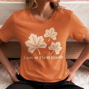 以下が含まれることがあります： オレンジ色のTシャツ。クリーム色のカエデの葉が3つと「Love at First Rustle」の文字がプリントされています。クルーネックで半袖です。デザインはTシャツの前面に配置されています。