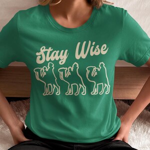 May include: A green t-shirt with the words "Stay Wise" in a cream-colored, stylized font. Below the text are three cream-colored camel silhouettes. The shirt is a crew neck and is being worn by a person.