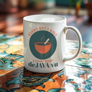 Könnte beinhalten: Weiße Keramik-Kaffeetasse mit gebogenem Henkel. Die Tasse trägt den Text "EVERY DAY I HAVE a Little bit of deJAVA vu" und eine Grafik einer dampfenden Schüssel und eines Löffels in einem tealfarbenen Kreis.