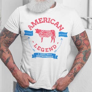 May include: White t-shirt with a red and blue graphic design. The graphic features a cow with a diagram of its cuts of meat, the words "American Legend", and the text "In Steak We Trust" and "A Real Beef Enthusiast".