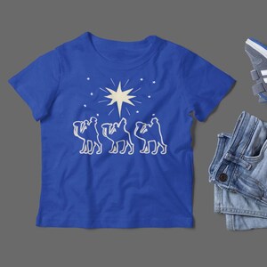 May include: A royal blue t-shirt with a white silhouette of three camels following a star. The star is a five-pointed star with a white outline and a yellow center.