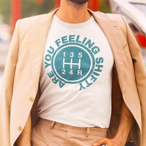 May include: A white t-shirt with a blue graphic of a car's gear shifter and the text "Are You Feeling Shifty".