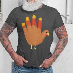 Puede incluir: Camiseta gris oscuro con un diseño de pavo dibujado a mano. El pavo está creado a partir de una forma de mano, con los dedos representando las plumas del pavo. Los dedos están coloreados en tonos naranja y amarillo, con pico y patas.