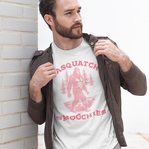 Peut inclure: Un t-shirt blanc avec un graphique rose d'un sasquatch debout dans une for&ecirc;t avec le texte "Sasquatch Smoochies".