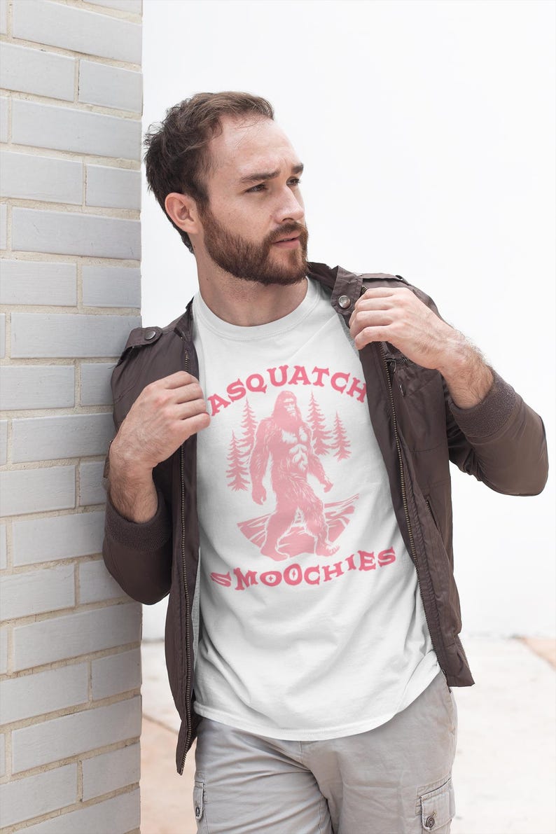 Peut inclure: Un t-shirt blanc avec un graphique rose d'un sasquatch debout dans une for&ecirc;t avec le texte "Sasquatch Smoochies".