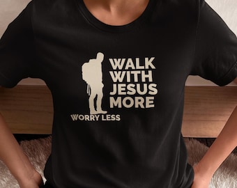 May include: Black t-shirt with the text "WALK WITH JESUS MORE WORRY LESS" in white. A white silhouette of a person with a backpack is on the left side of the text. The shirt is made of a soft material.