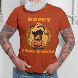 Pode incluir: Uma camiseta laranja queimada com um gato preto sentado em cima de uma abóbora esculpida. O texto "HAPPY YOWL-O-WEEN" está impresso na camiseta.