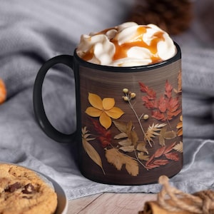 May include: Black coffee mug with a brown woodgrain design featuring pressed autumn leaves in shades of yellow, orange, and red.
