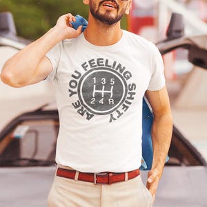 May include: White t-shirt with a black and gray graphic of a car's gear shifter with the text "Are you feeling shifty?" around it.