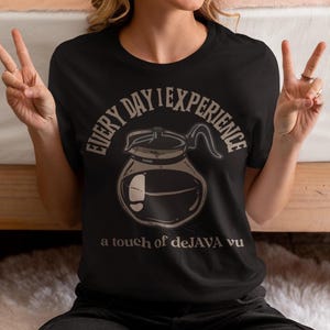 Peut inclure: T-shirt noir avec un dessin de cafetière et le texte "EVERY DAY I EXPERIENCE a touch of deJAVA vu" en marron clair. Le dessin de la cafetière est un simple dessin au trait. Le t-shirt est de style décontracté.