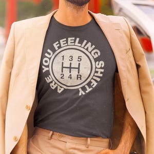May include: A dark gray t-shirt with a white graphic of a car's gear shifter and the text "Are you feeling shifty?"