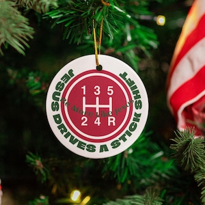 May include: A round, white Christmas ornament with a red and green gear shift pattern. The ornament reads "Jesus Drives a Stick Shift" in green text.