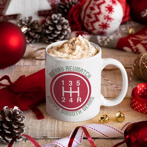 Könnte beinhalten: Weiße Keramik-Kaffeetasse mit einem rot-weißen Weihnachtsmotiv. Die Tasse zeigt einen roten Kreis mit einem weißen Schaltmuster und dem Text "Merry Reumatch Happy Smooth Gears".