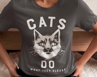 May include: Dark grey t-shirt featuring a white graphic of a cat's face and the text "CATS DO WHAT THEY PLEASE". The cat illustration has detailed whiskers and a stylized design. The t-shirt is a casual wear item.
