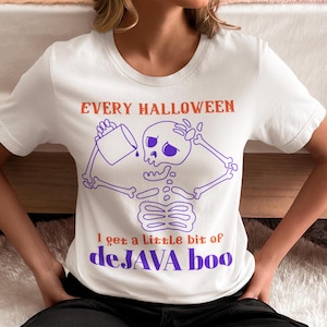 Op de afbeelding: Wit T-shirt met een paarse skeletafbeelding die uit een mok giet. De tekst luidt "EVERY HALLOWEEN" in oranje en "I get a little bit of deJAVA boo" in paars en oranje. Een leuke Halloween-thema tee.